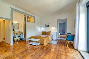 Une pièce avec un plancher en bois, un lit recouvert d’une couvre-lit beige, une table de chevet blanche, un bureau avec une chaise et une porte donnant sur une autre pièce.