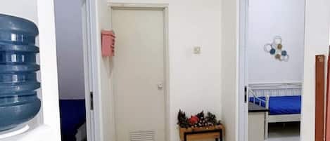 Un pasillo estrecho que conduce a una habitación con cama, una mesita con adornos y un dispensador de agua.