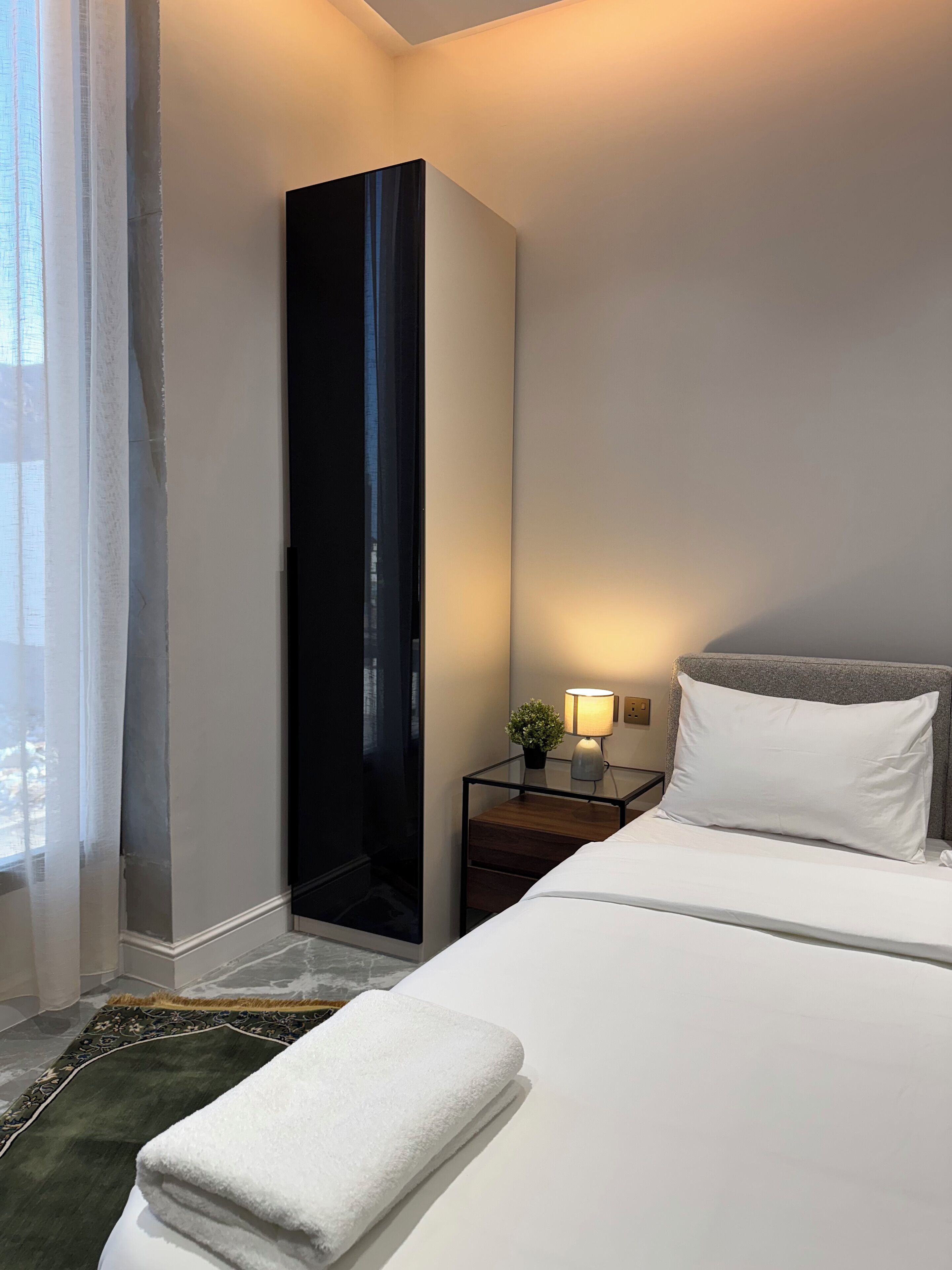 A hotel room with a bed, a nightstand with a lamp, a potted plant, and a large window with curtains.