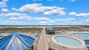 Een houten terras met lounge stoelen en een gazebo met uitzicht op een strand en een heldere lucht.