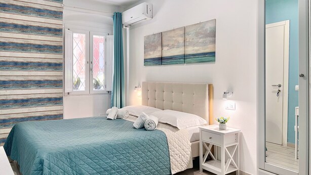 A bedroom with a bed, a bedside table with a lamp, a window with curtains, and a wall-mounted air conditioner. - La Dolce Cagliari Holiday Home (Cagliari)