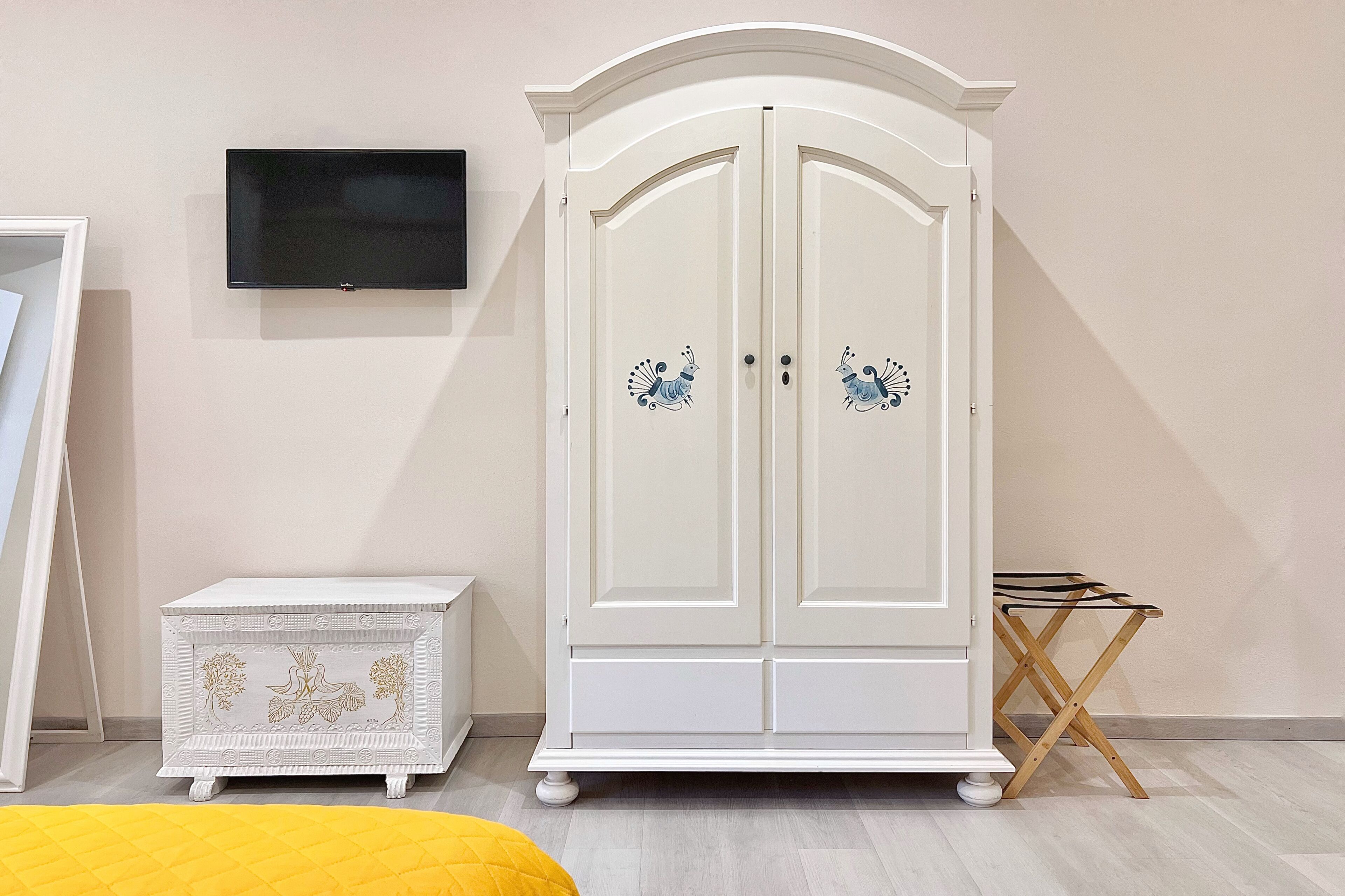 Ein Zimmer mit einem weißen Kleiderschrank, einer kleinen weißen Kommode, einem an der Wand befestigten Flachbildfernseher und einem hölzernen Klappstuhl.
