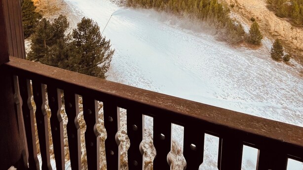 Un balcón con vista a un paisaje nevado, que incluye un río, un bosque y un telesilla.