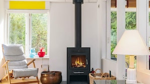 A cozy living room with a wood-burning stove, a striped chair, a table, and a view of greenery outside. - 6 Person Holiday Home in Stege (Stege)