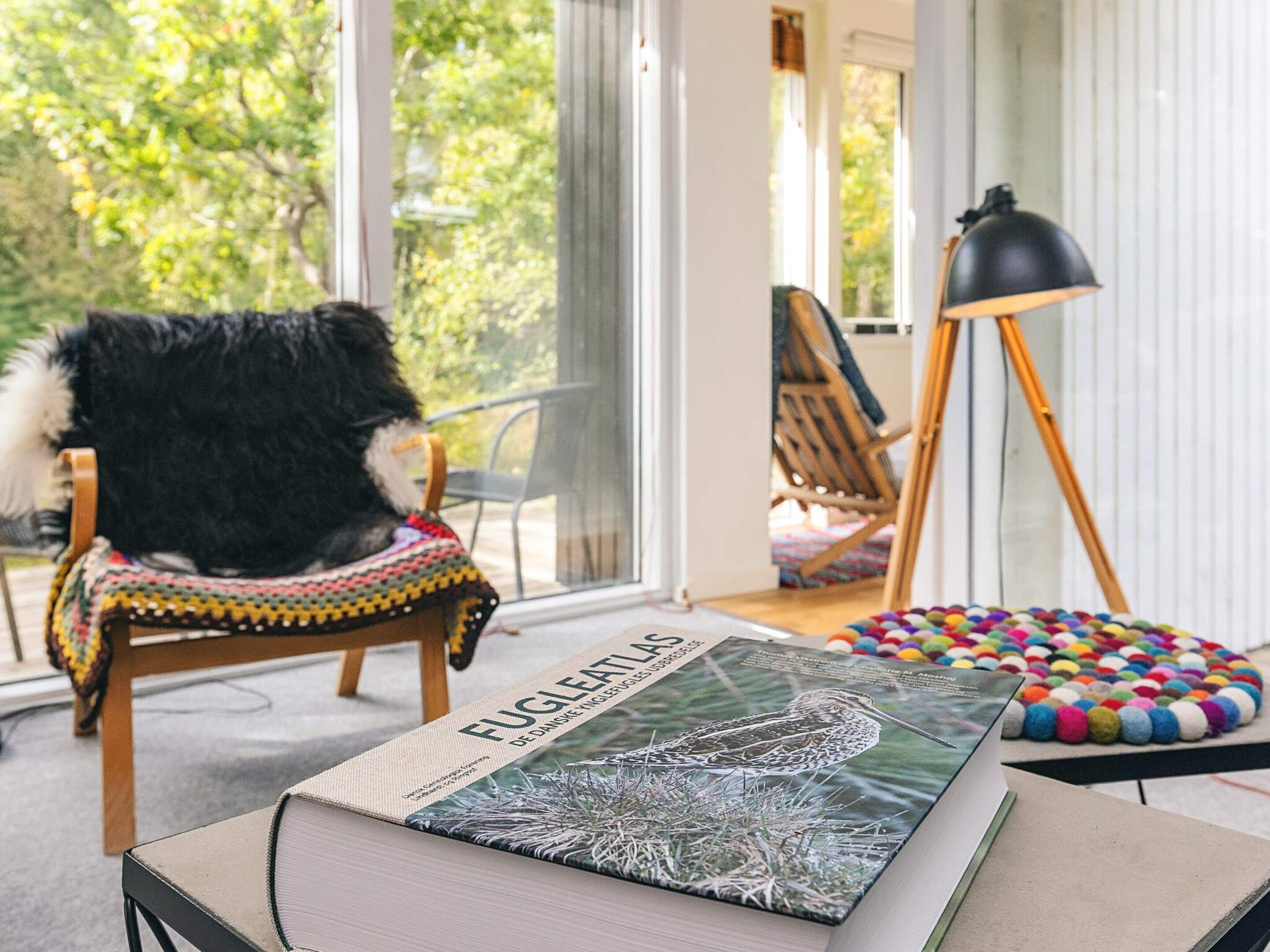 A room with a large window, a chair with a fur throw, an easel, a book titled 'Fugletti,' and a colorful pom-pom rug.