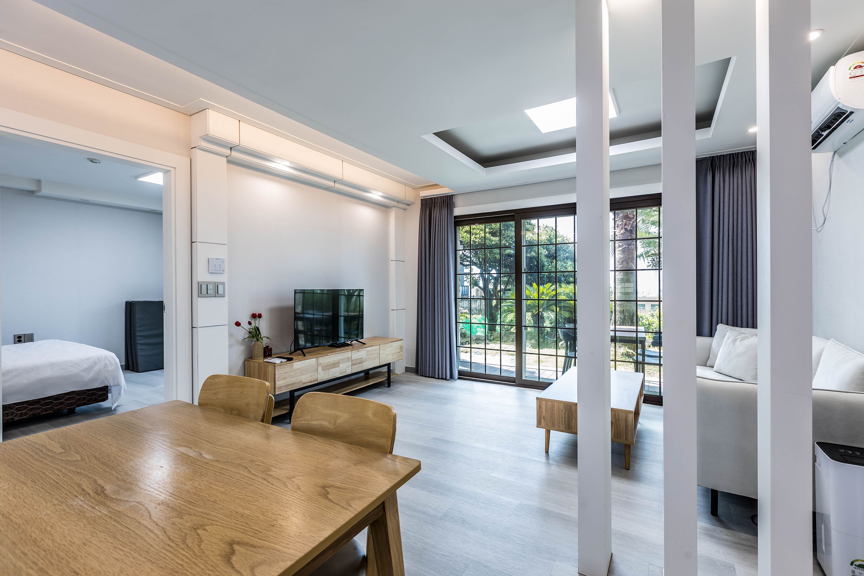 A modern hotel room with a wooden dining table, a sofa, a TV, and a large window with curtains.