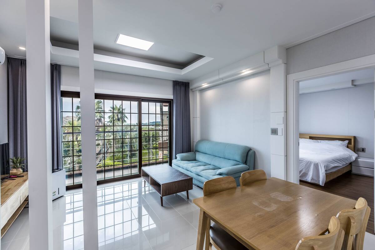Una habitación de hotel moderna con una mesa de comedor de madera, un sofá azul, una cama con ropa de cama blanca y un ventanal con vistas a zonas verdes.