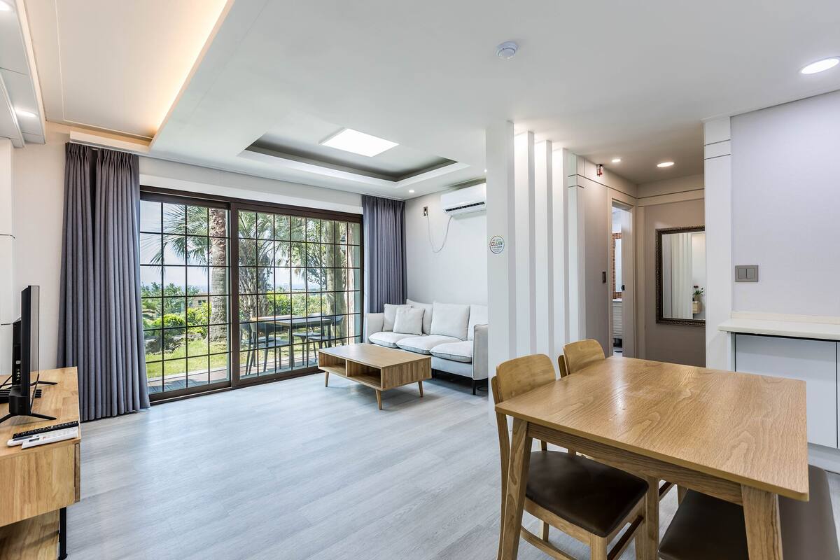Una habitación de hotel moderna con un ventanal grande, un sofá, una mesa de comedor de madera y un televisor.