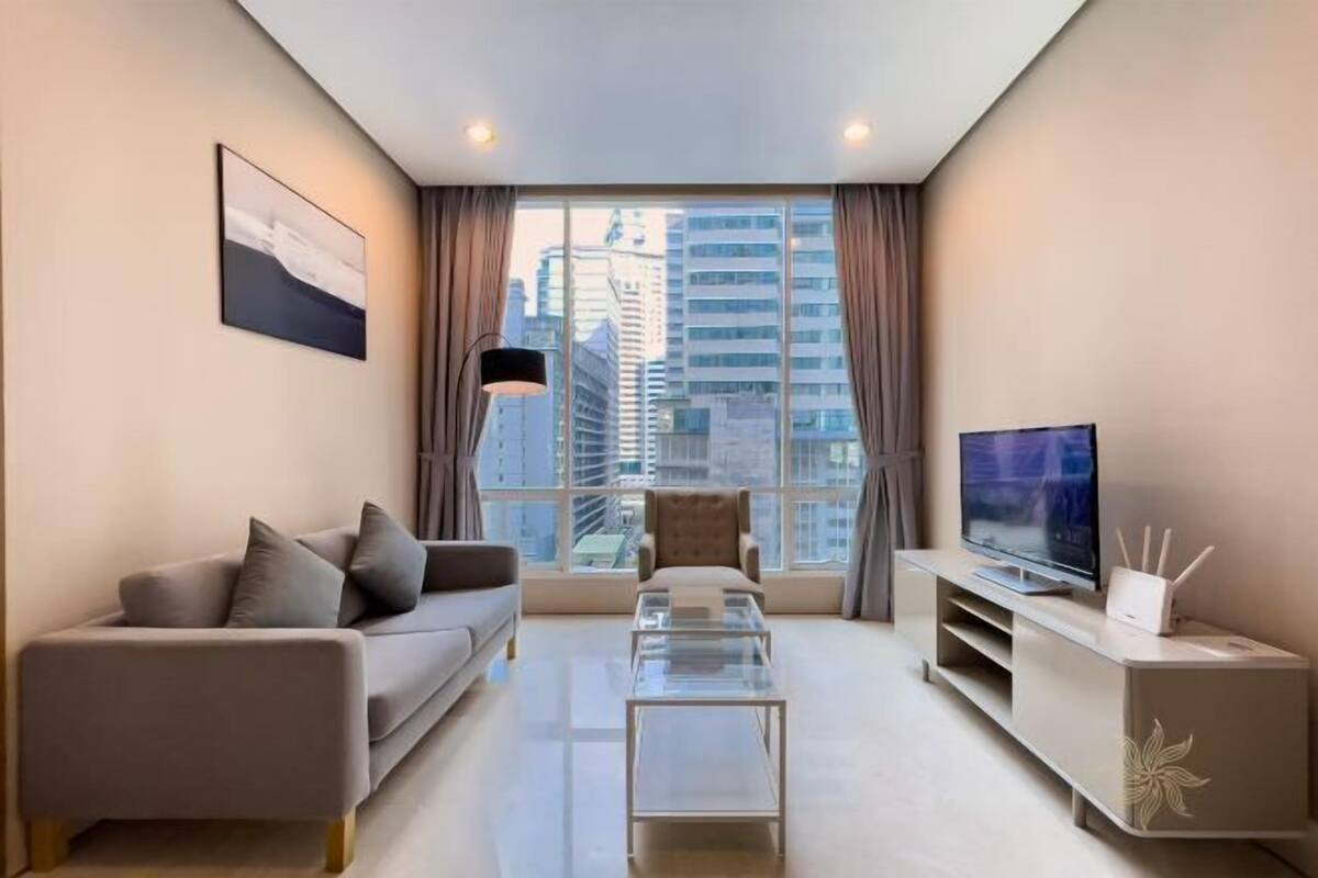 A modern living room with a sofa, a glass coffee table, a TV, and a large window with curtains.