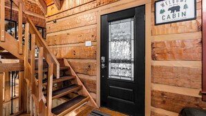 A wooden cabin interior with a black door, a welcome sign, and a wooden staircase. - Smoky Mountain Views | Hot Tub | Game Room | 4BR (Gatlinburg)