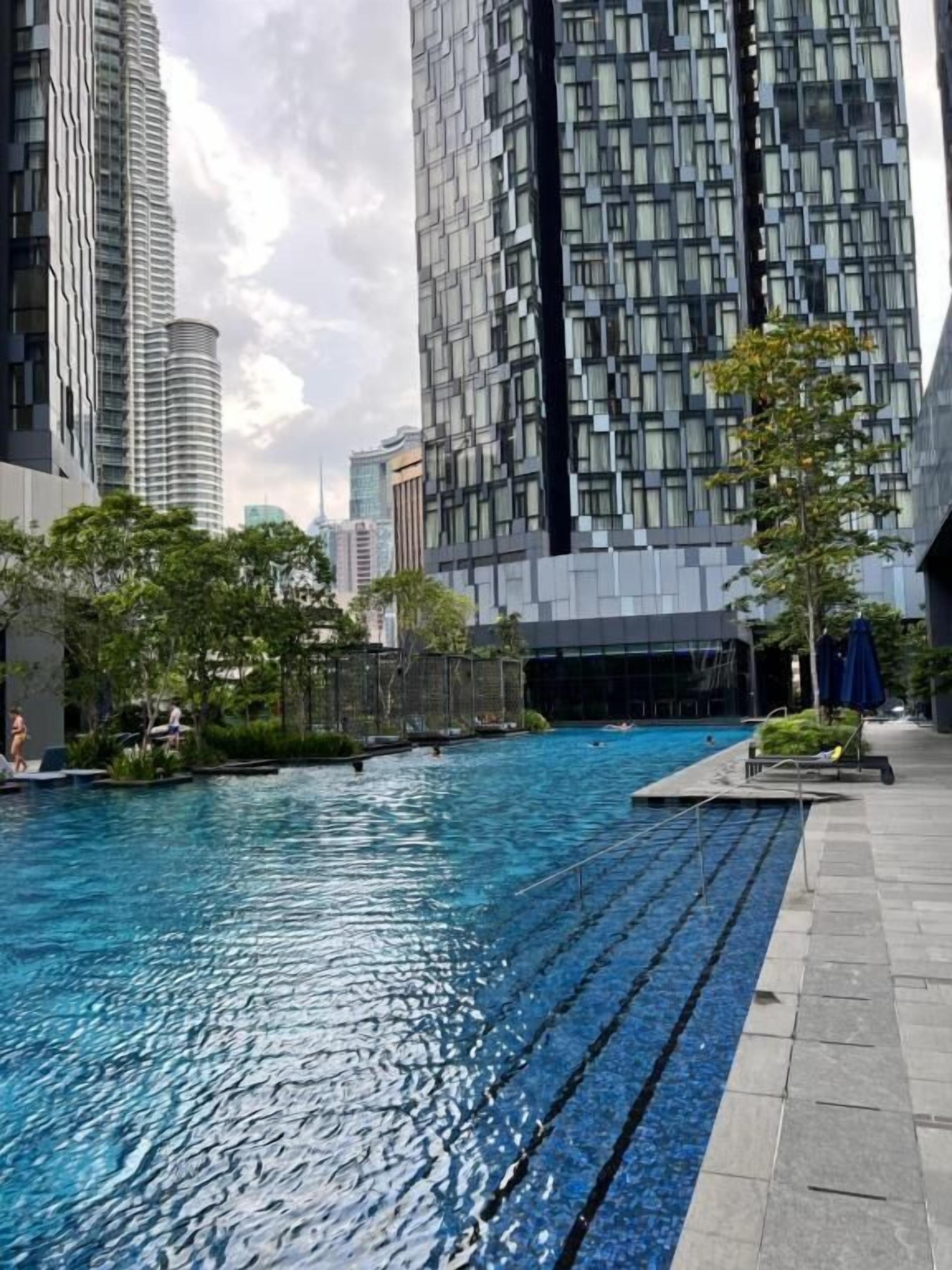 Une piscine dont le bord est revêtu de carrelage, entourée de verdure et de bâtiments modernes.