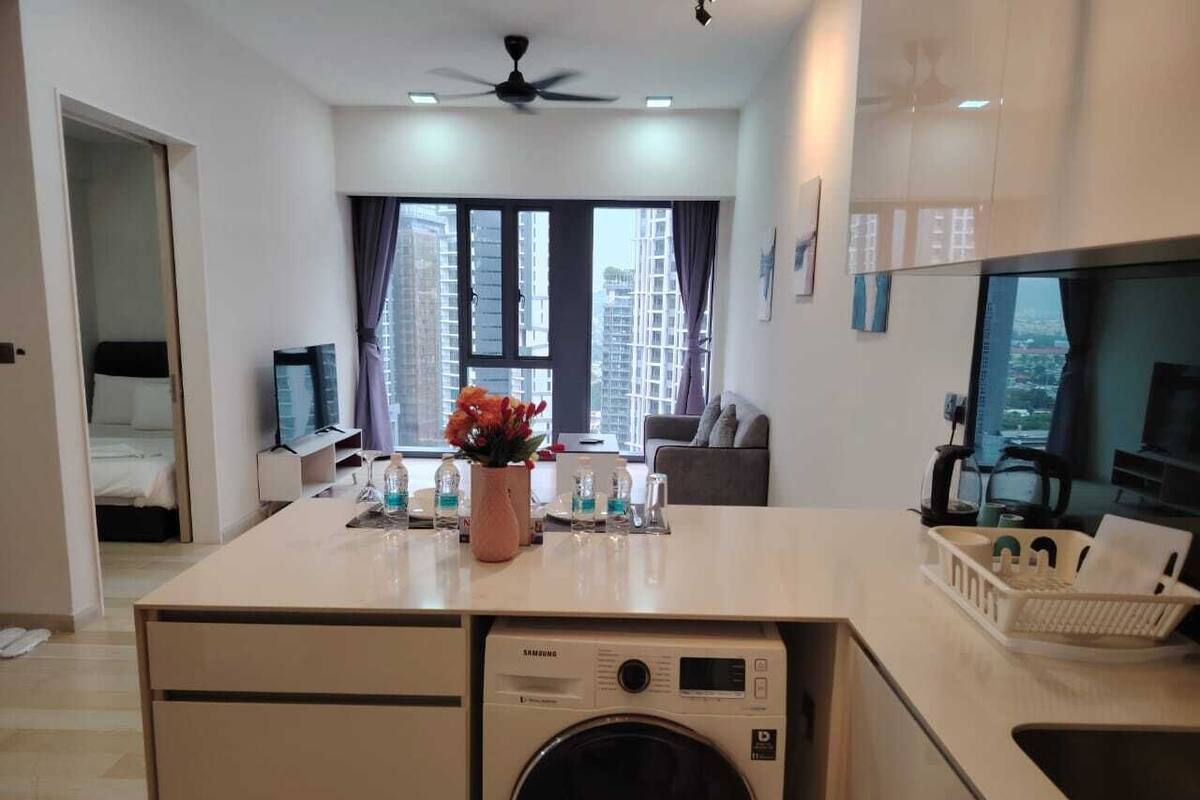 A modern apartment with a central kitchen area featuring a Samsung washing machine, a dining area with a view of high-rise buildings, and a living space with a sofa and a TV.