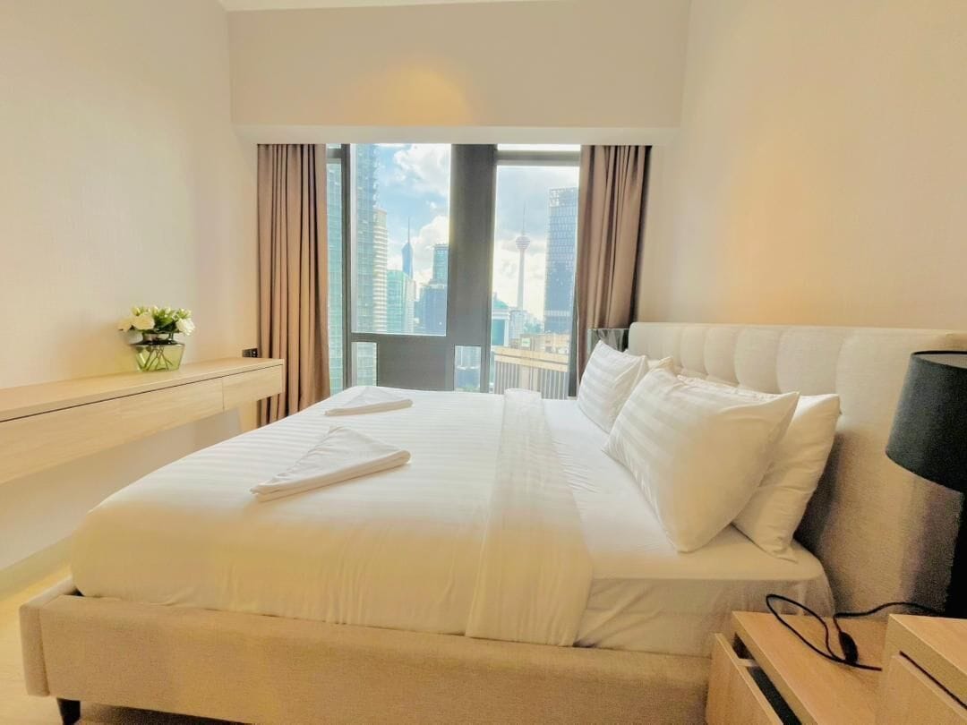 Camera d'albergo con un letto grande, vista sulla città e un tavolino con una lampada.