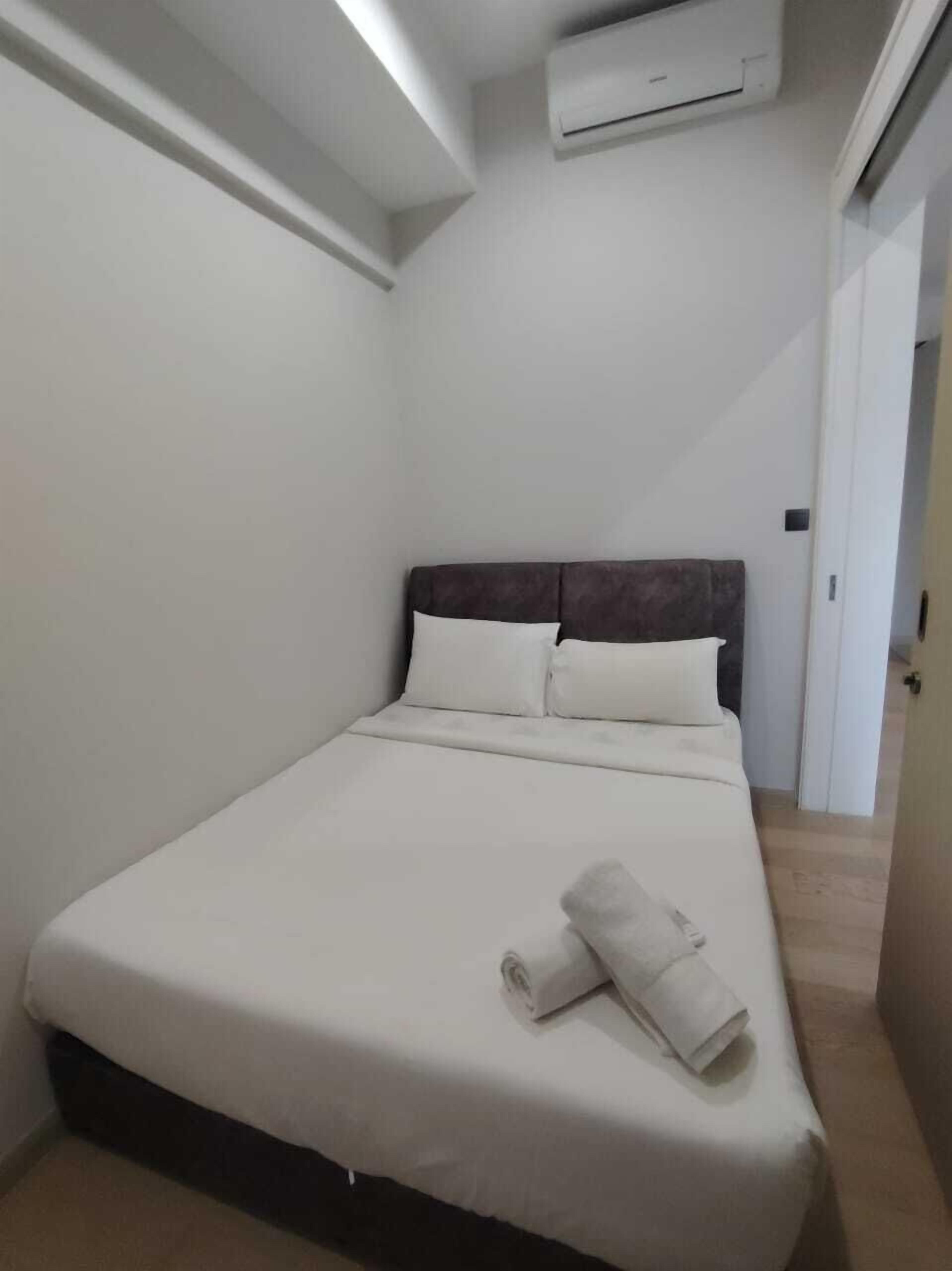 Een klein, net opgemaakt bed met witte lakens en kussens, een airconditioning erboven en een deur aan de rechterkant.