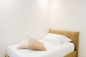A bed with white bedding and a tan headboard in a room with white walls.