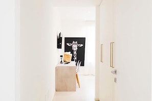 A hallway with a minimalist design, featuring a white door with gold handles, a black and white artwork on the wall, and a small table with a chair.