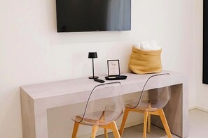 Una habitación moderna con un televisor de pantalla plana montado en la pared, una consola blanca con mando a distancia, un cuadro enmarcado y un jarrón decorativo. Dos sillas transparentes con patas de madera están colocadas frente a la consola.