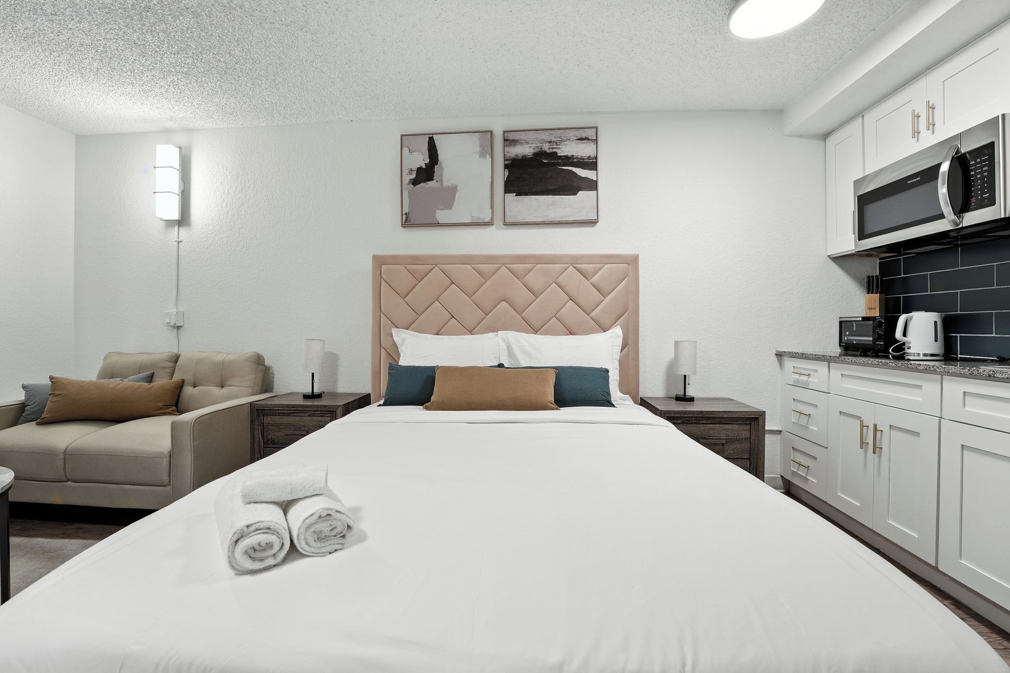 Quarto de hotel com uma cama grande, um sofá, uma cozinha compacta e duas obras de arte na parede.