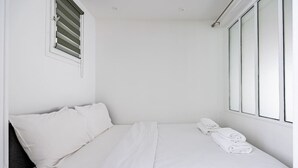A small, white, single-bed room with a window and a wall-mounted air vent.