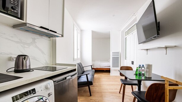 A compact kitchen with a washing machine, a kettle, and a microwave. There is a dining area with a table and chairs, and a bed in the background.