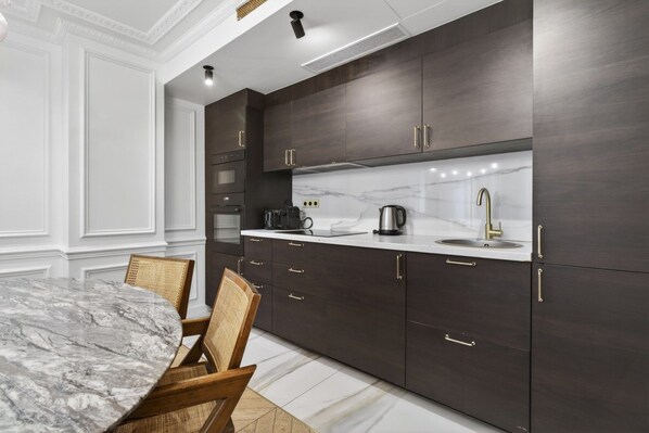 A modern kitchen with dark cabinetry, a marble countertop, and a dining area with a marble table and wicker chairs.