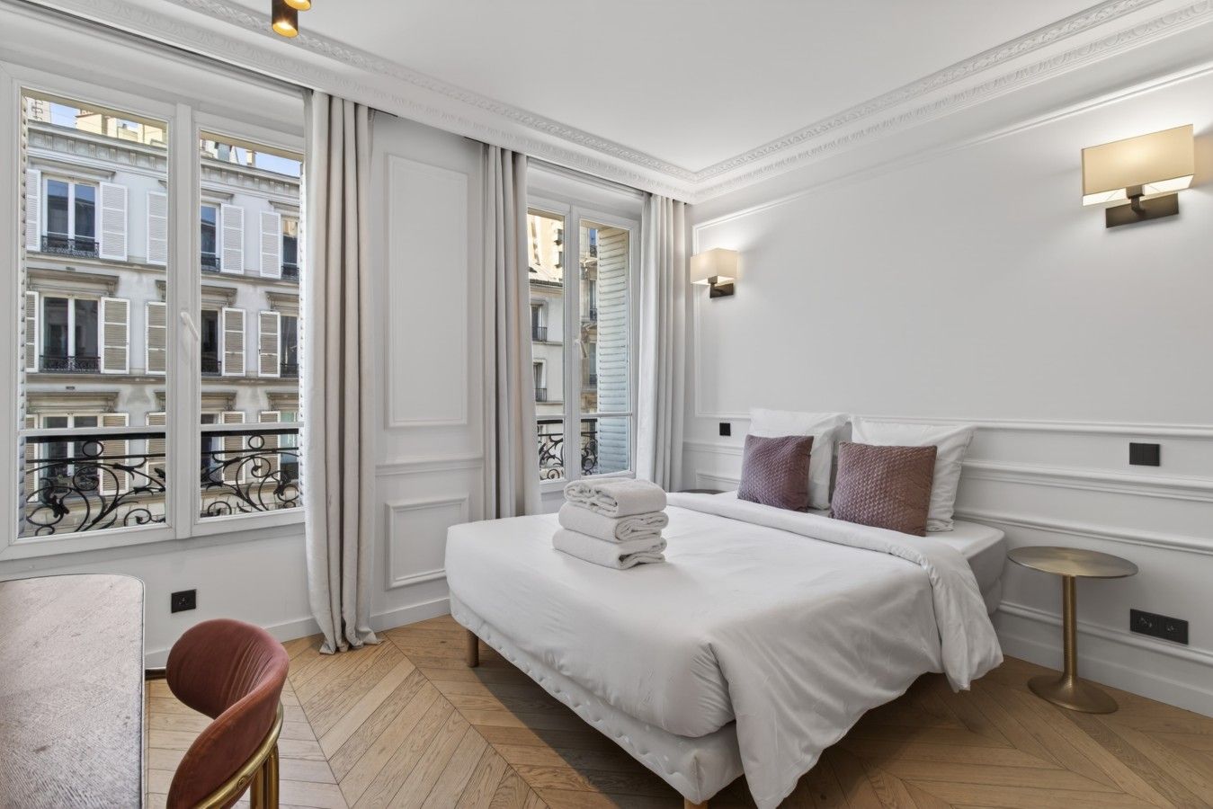 A hotel room with a large bed, a small round table, a chair, and a view of the city through the windows.