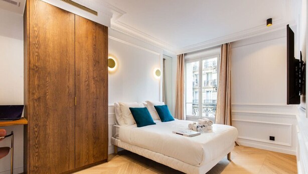 A modern hotel room with a wooden wardrobe, a bed with blue pillows, a desk with a laptop, and a view of the city through large windows. - #3Bedrooms#Lafayette#Paris10#AC (Paris)