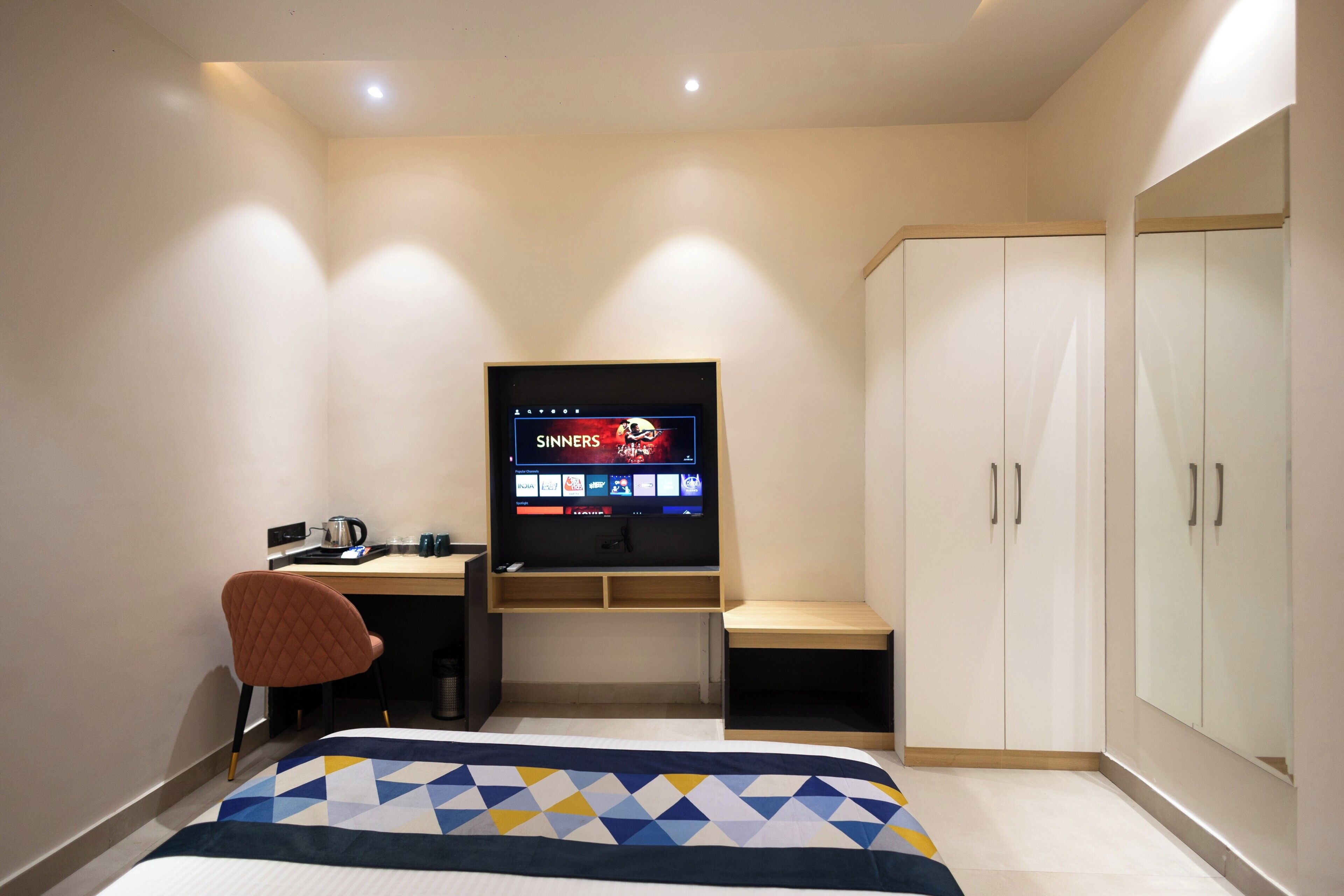 Een slaapkamer met een bed, een bureau met een computer, een stoel, een kledingkast en een televisietoestel dat een programma toont.