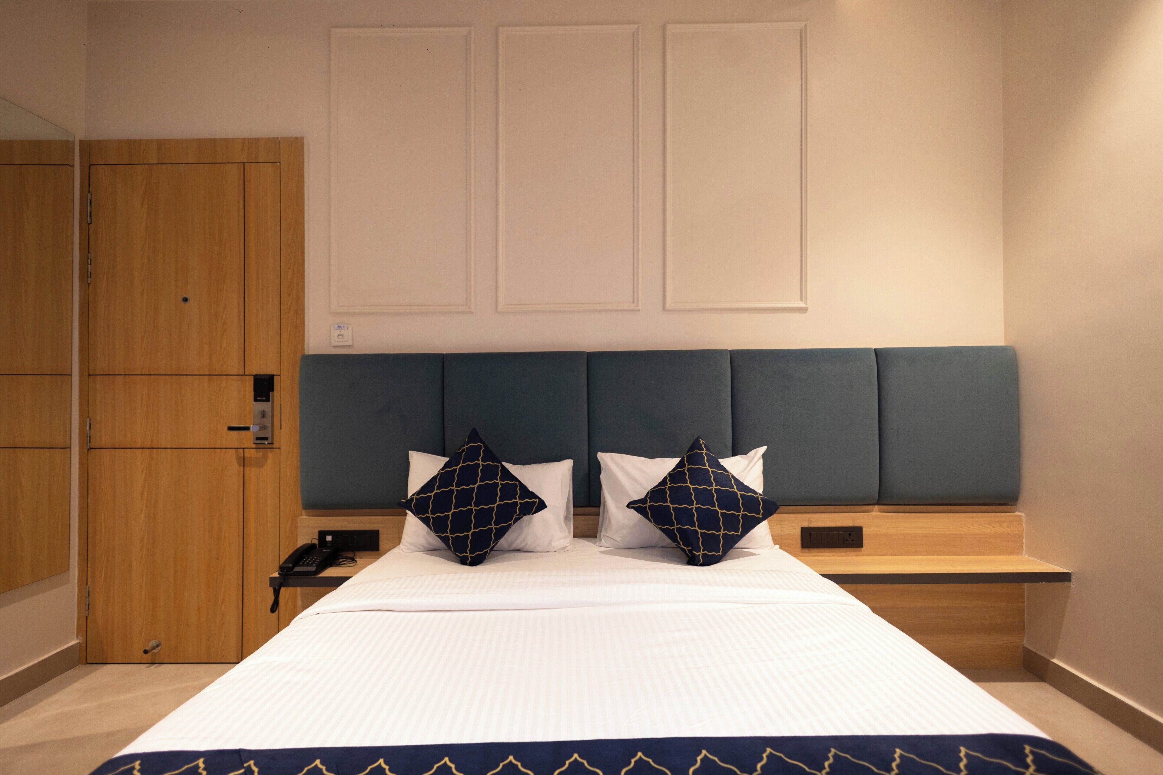 Una habitación de hotel con cama, dos almohadas, una mesita de noche, un armario de madera y un panel en la pared.