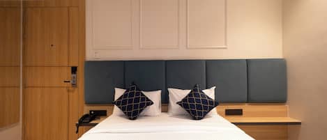 A hotel room with a bed, two pillows, a bedside table, a nightstand, a wooden wardrobe, and a wall-mounted panel.