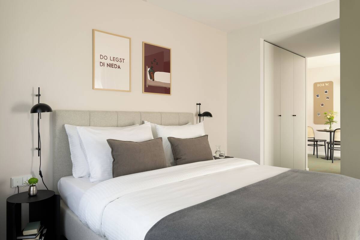A neatly made bed with white and gray bedding, a small bedside table with a lamp, and a wall-mounted picture frame with text.