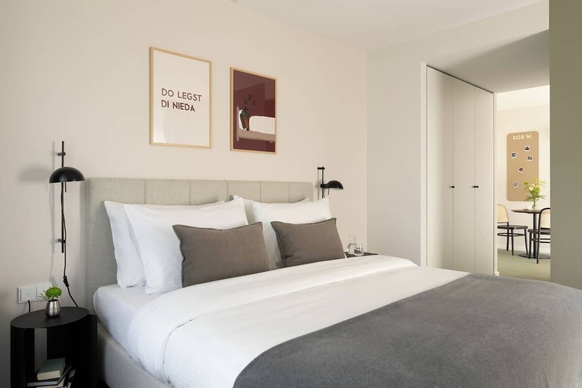 Un lit bien fait, avec une literie blanche et grise, une petite table de chevet avec une lampe et un cadre photo fixé au mur, sur lequel on peut voir du texte.