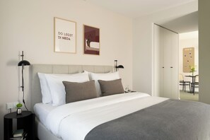 A neatly made bed with white and gray bedding, a small bedside table with a lamp, and a wall-mounted picture frame with text. - Bob W Munich City West (Munich)
