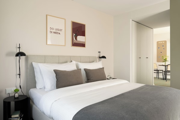 Una cama bien hecha con ropa de cama blanca y gris, una mesita de noche con una lámpara y un cuadro con texto en la pared.