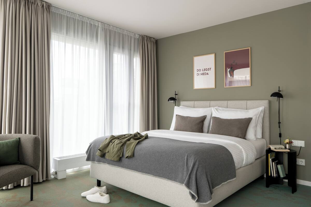 A bedroom with a bed, a chair, a bedside table with books, and a window with curtains.