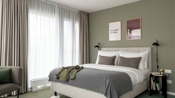 Un dormitorio con una cama, una silla, una mesita de noche con libros y una ventana con cortinas.