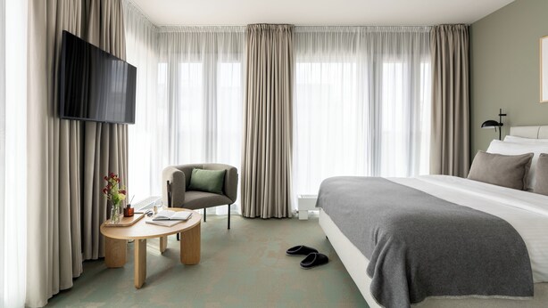 Een hotelkamer met een bed, een televisie, een stoel, een tafel met een boek en een vaas, en gordijnen.