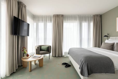 Ein Hotelzimmer mit Bett, Fernseher, Sessel, Tisch mit Buch und Blumenvase, sowie Vorhängen.. Bob W Munich City West