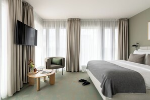 Habitación de hotel con cama, televisor, silla, mesa con un libro y un jarrón, y cortinas.