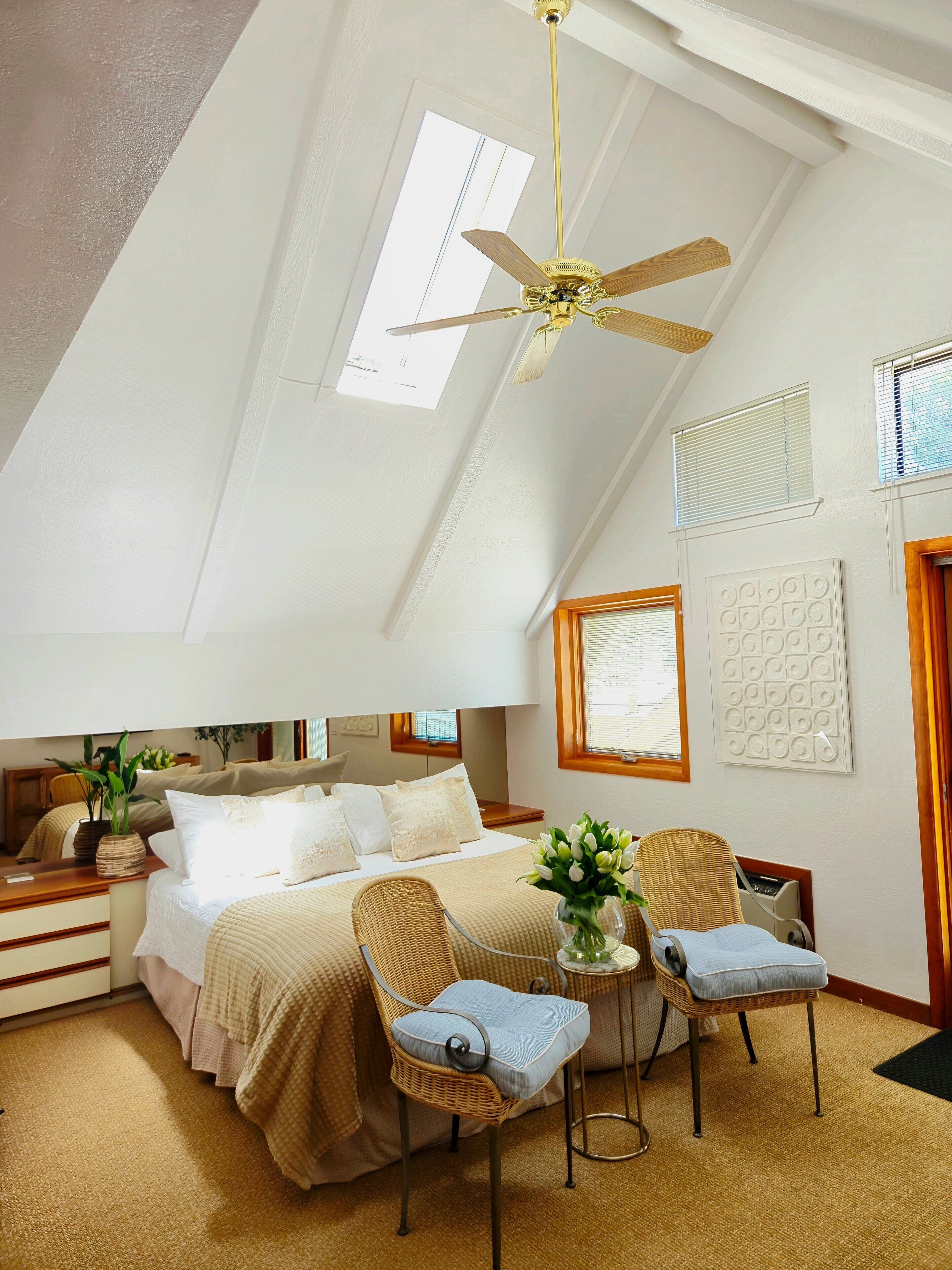A bedroom with a bed, two chairs, a small table, a ceiling fan, and a window.