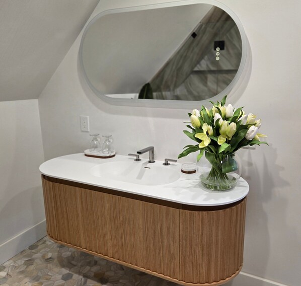 Une salle de bain avec un meuble-lavabo en bois, un lavabo blanc, un miroir et un vase de fleurs.