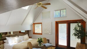 A bedroom with a sloped ceiling, a bed with white linens, a wicker chair, a small table with a vase, and a wooden door. - Tahoe Vista Inn Suite 5 (Tahoe Vista)