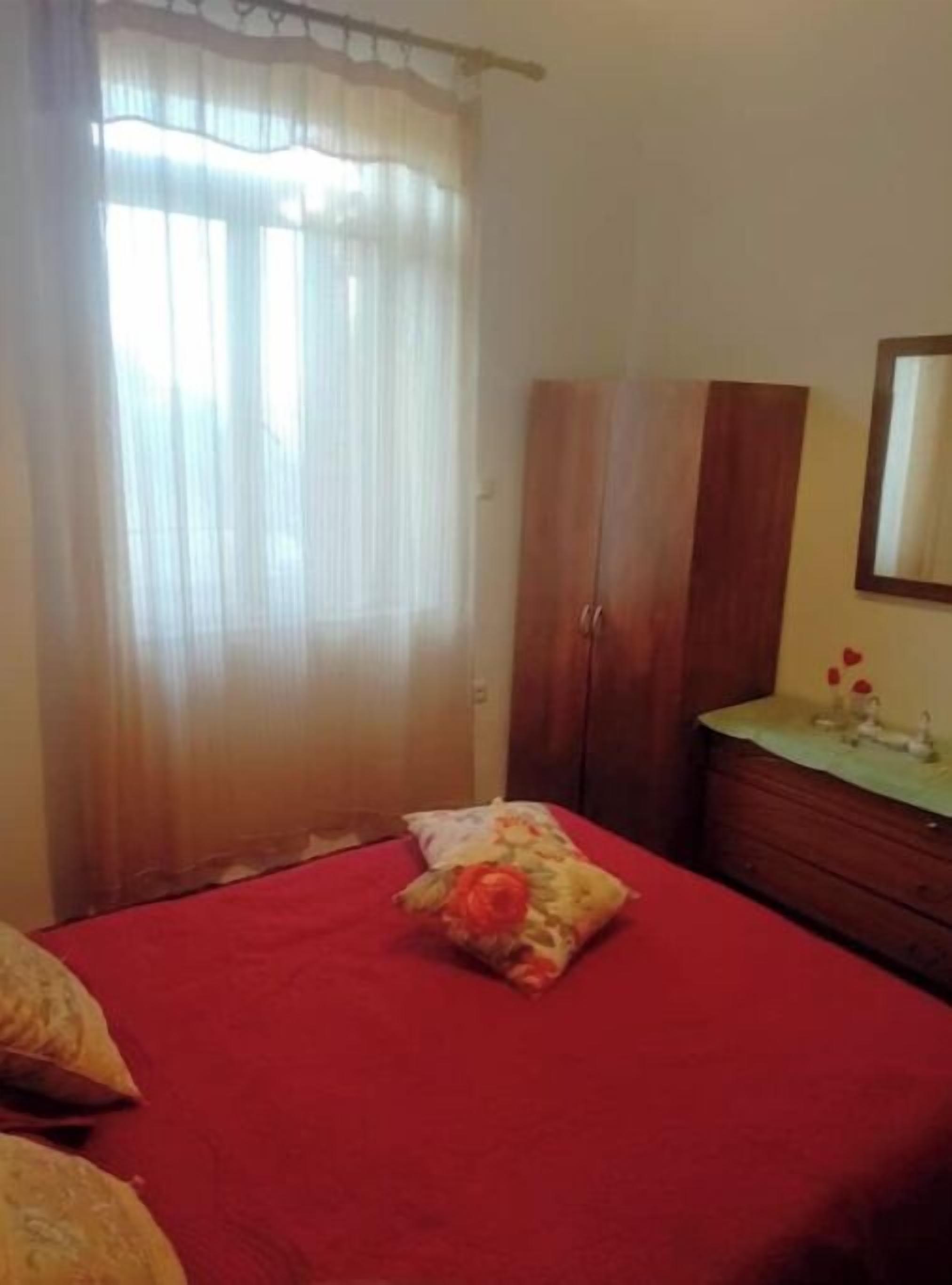 A bedroom with a red bedspread, a wooden wardrobe, a bedside table with a mirror, and sheer curtains on the window.