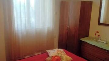 A bedroom with a red bedspread, a wooden wardrobe, a bedside table with a mirror, and sheer curtains on the window.
