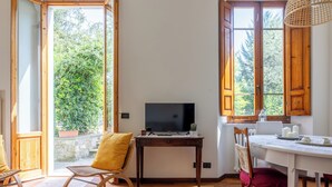 Ein Zimmer mit Schreibtisch aus Holz, einem Fernseher und einem Esstisch mit Stühlen. Große Fenster mit Holzläden bieten Blick ins Grüne.