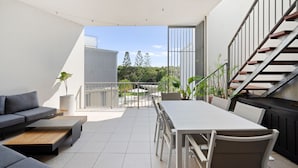 Un espacio exterior moderno con sofá, mesa de comedor y sillas, una escalera y vistas al jardín y árboles.