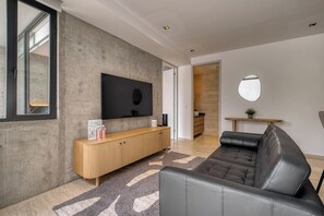 A modern living room with a leather sofa, a wooden console table, and a flat-screen TV mounted on a concrete wall.