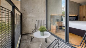 Uma varanda moderna com uma mesa pequena, cadeiras, um vaso de planta e vista para um quarto com cama e cabeceira estampada.