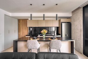 A modern kitchen with a central island, two bar stools, and a dining area with a black leather couch.