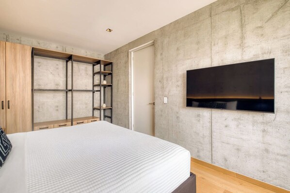 A bedroom with a large bed, built-in wooden wardrobe, a flat-screen TV mounted on the wall, and a concrete-textured wall.